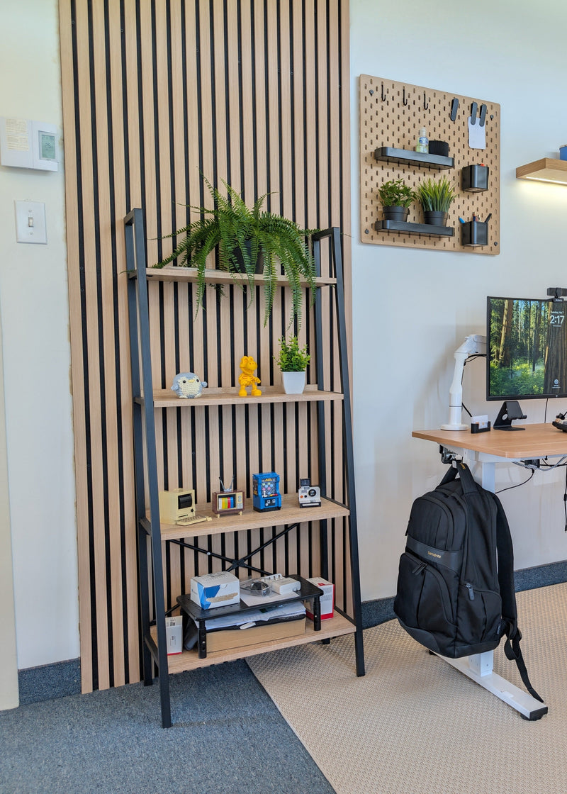 A modern home office setup featuring a standing desk with dual computer monitors, a black ergonomic office chair, a black backpack hanging from the desk frame, and a tall floor lamp. The room has white walls, a slatted wood accent panel on the left, and a beige rug.