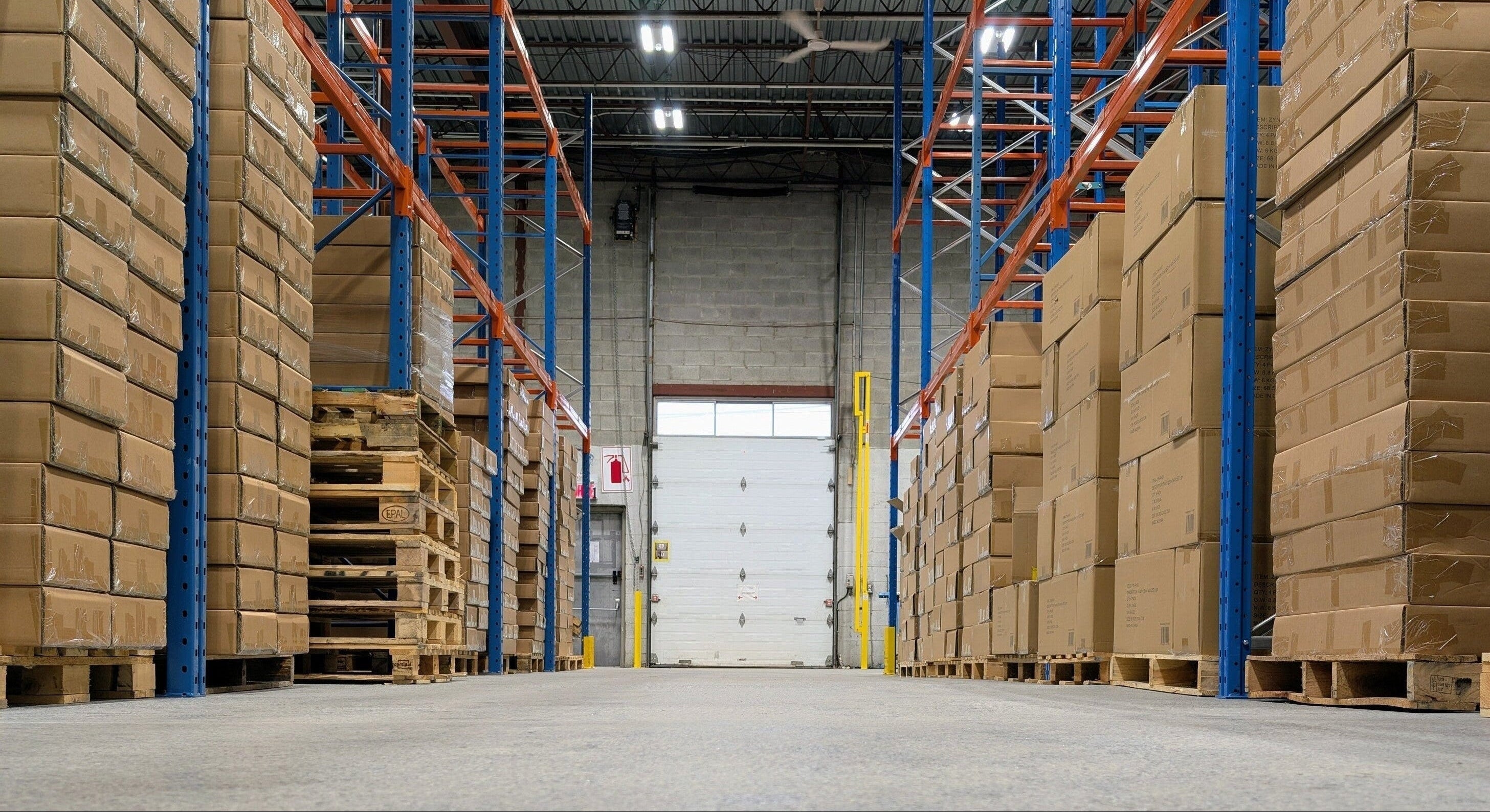 Warehouse interior with stacked cardboard boxes on pallets and high racking systems.