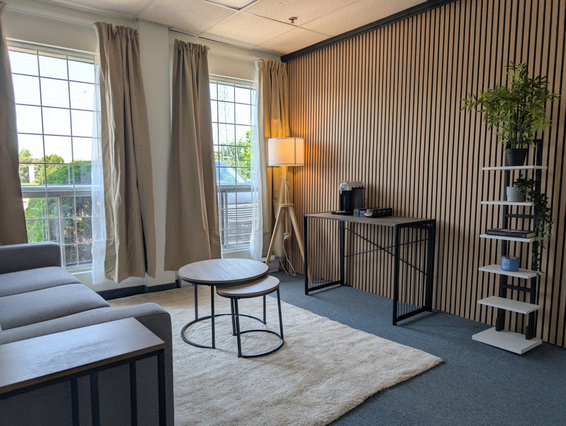 A modern living room interior with a gray sofa, round nesting tables on a white rug, a floor lamp, a slatted wood accent wall, and a white shelving unit with plants.