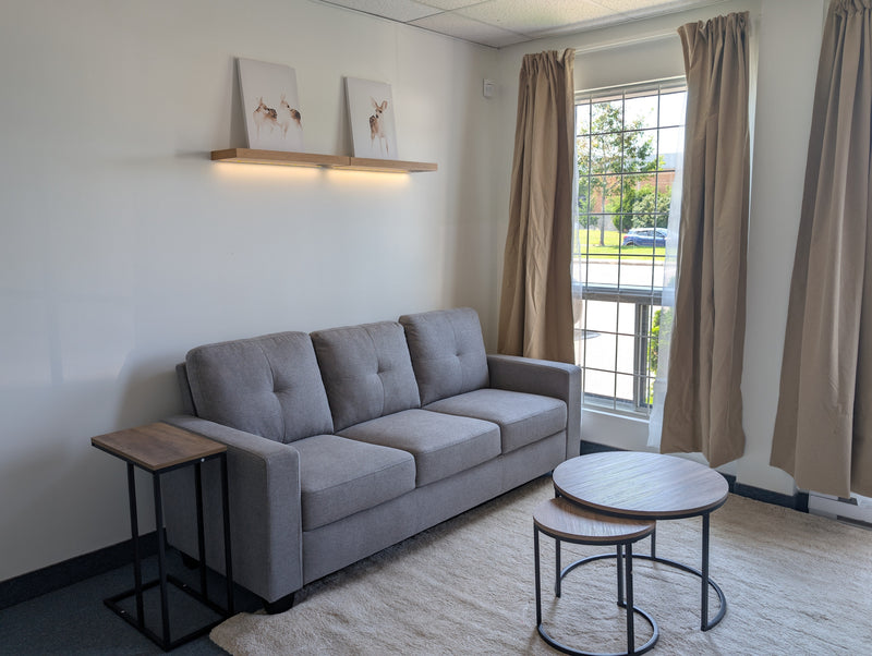 A modern gray fabric three-seater sofa in a bright, minimalist living room with beige curtains, a small wooden side table, and two round nesting coffee tables.