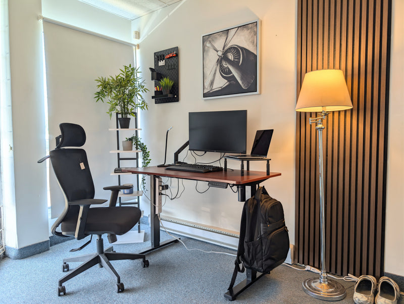 A modern home office setup featuring a wooden height-adjustable standing desk with a black computer monitor, keyboard, and laptop. A black ergonomic office chair is positioned to the left, and a black backpack hangs from the desk leg.