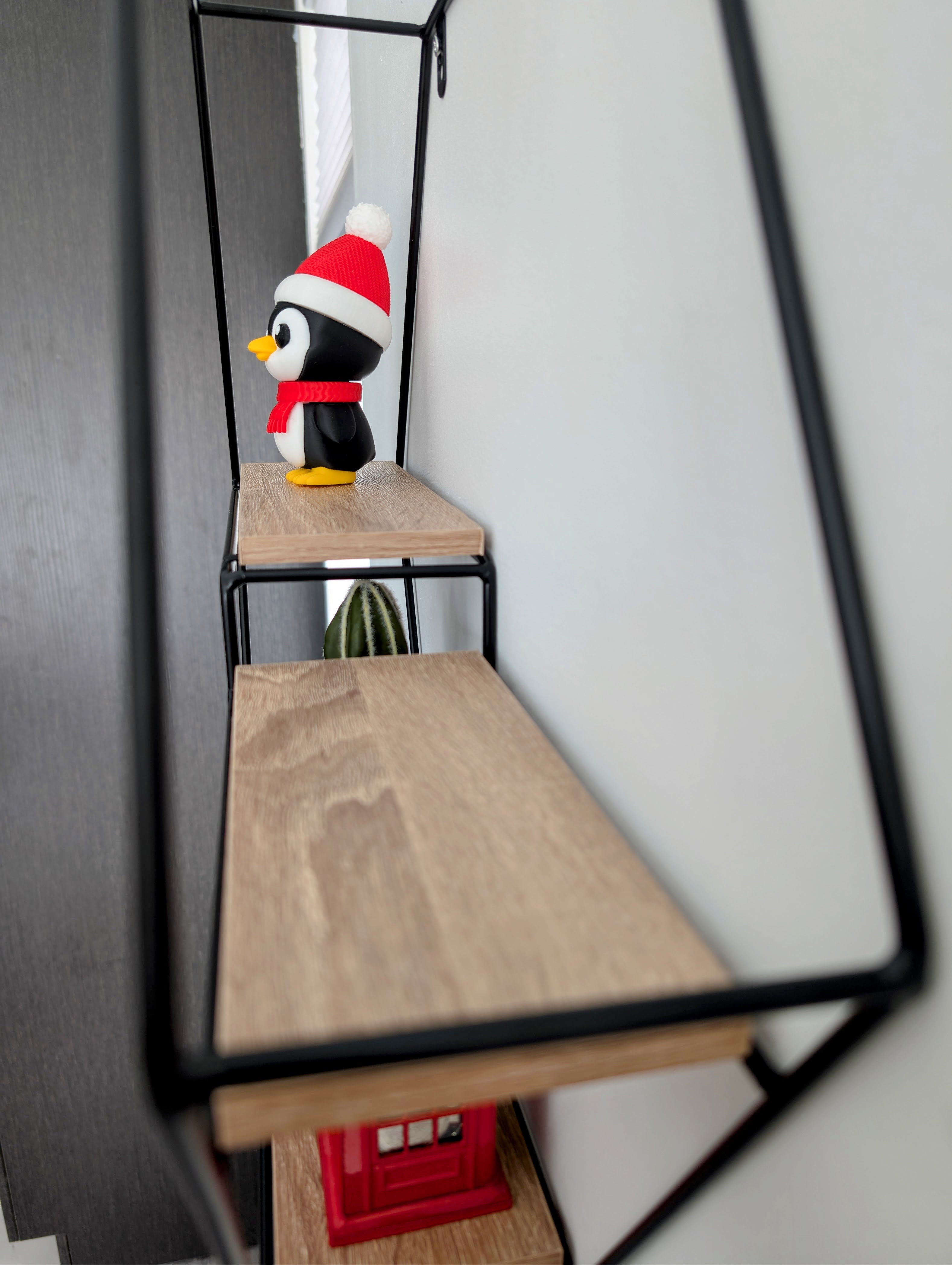 Small penguin figurine wearing a Santa hat on a wooden shelf inside a black frame.