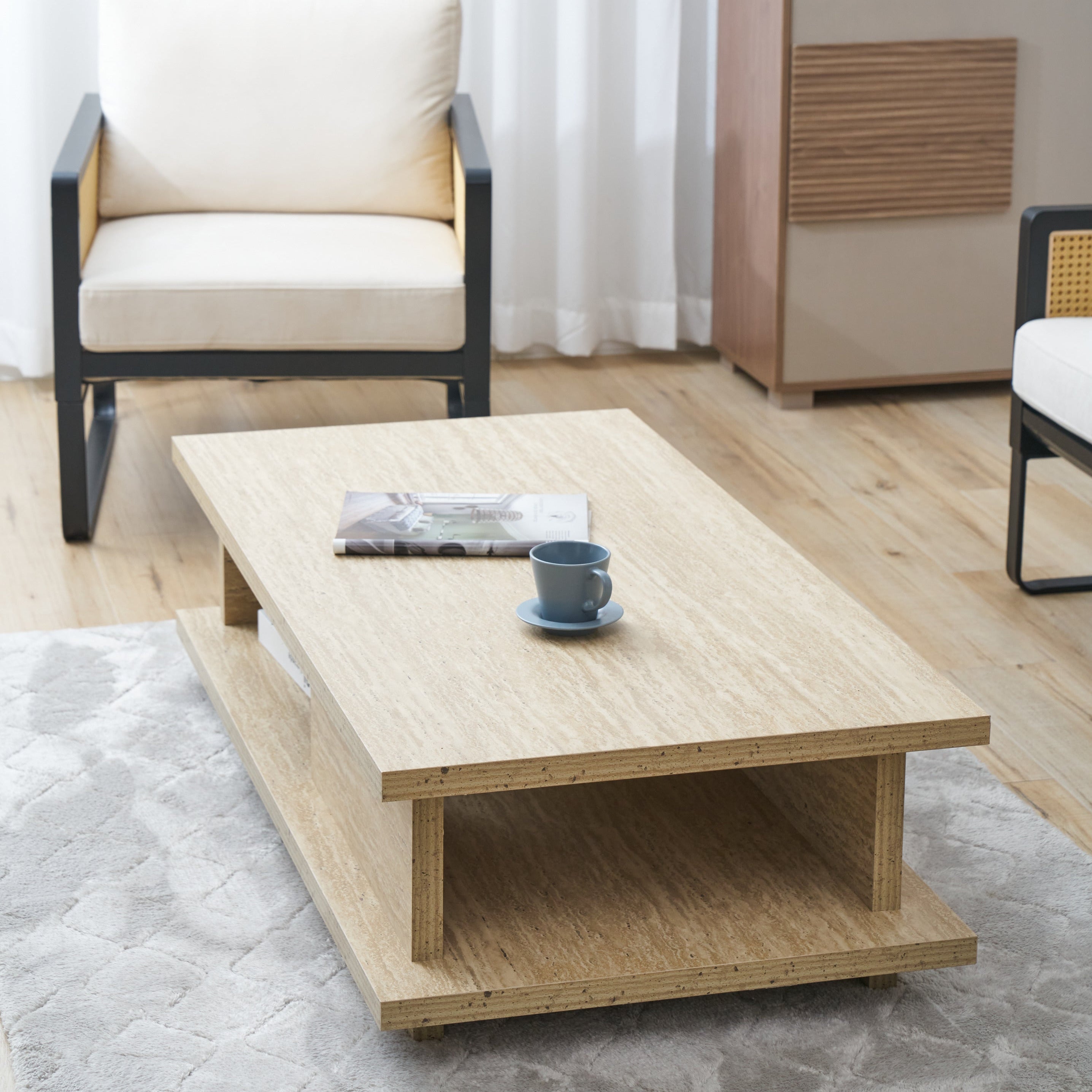 A light natural wood rectangular coffee table with an open lower shelf, styled in a bright living room with a gray rug, a cream armchair, and a wooden cabinet in the background.