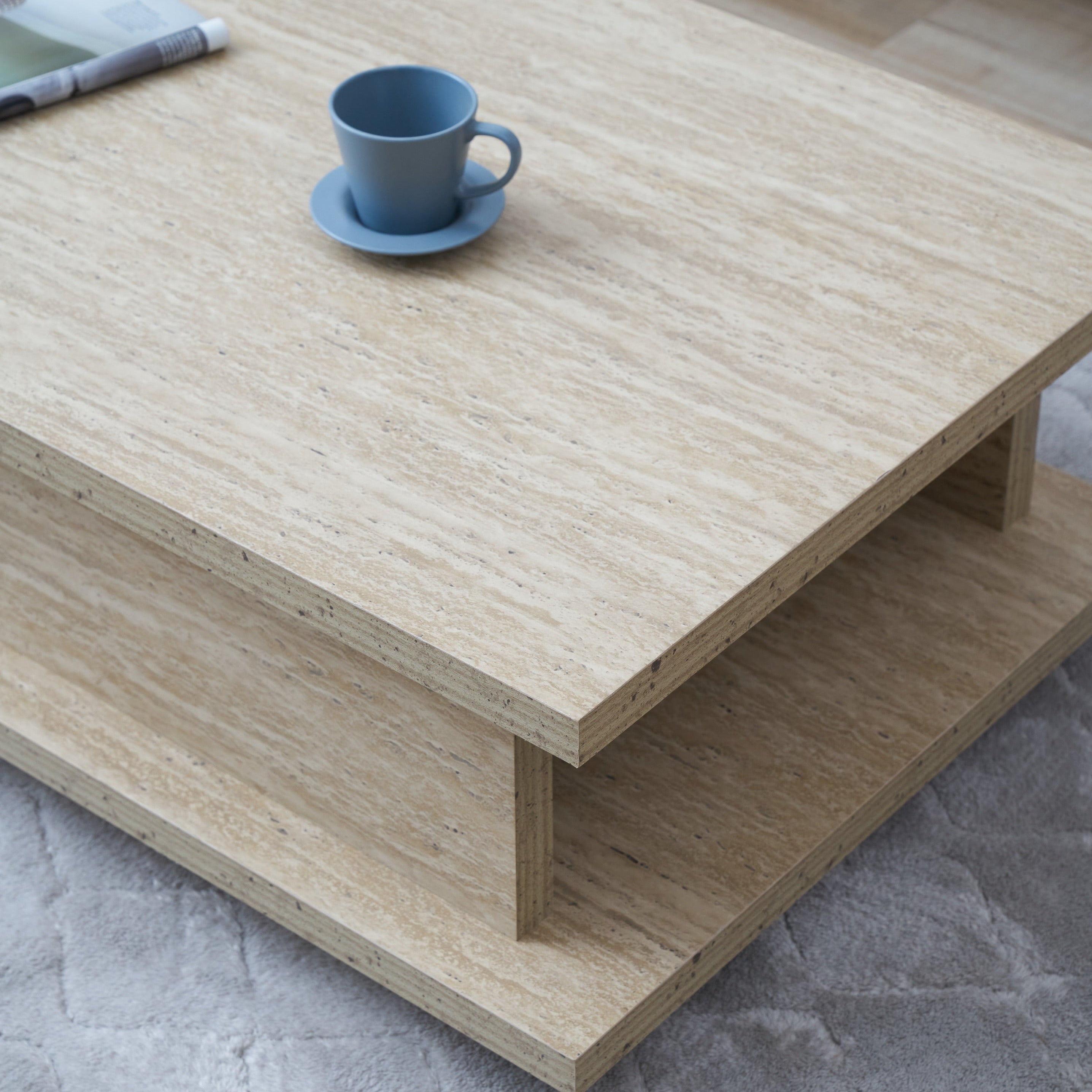 A light natural wood low coffee table with a double-tiered design, styled on a gray textured rug with a blue cup on top.